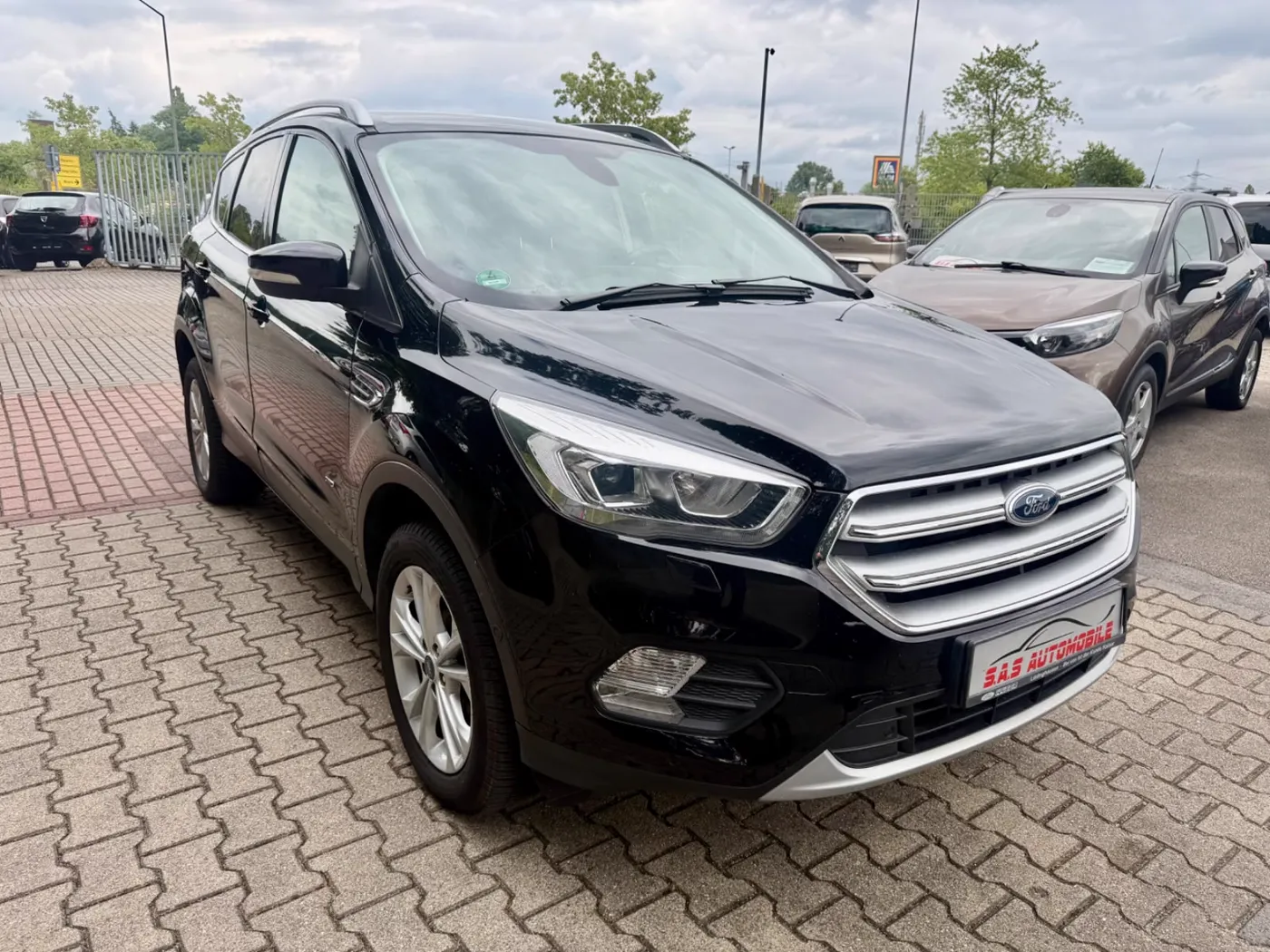 Schwarzer Ford Kuga Titanium auf dem gepflasterten Hof von S.A.S Automobile, dahinter weitere Gebrauchtwagen